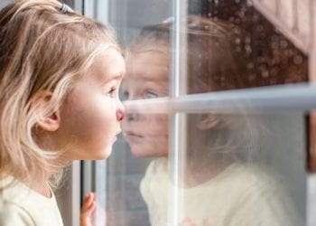 Little adorable blonde toddler girl looking through a window with rain drops on it. Close up portrait