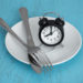 Intermittent fasting concept with clock on white plate, fork and knife on blue table