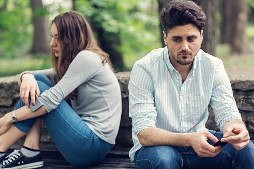 Young sad looking couple sitting at a distance and looking away from each other. Selective focus on the male.