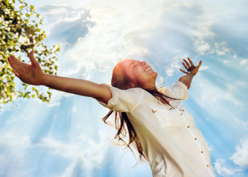 Pretty young woman raising her arms and enjoying perfect sunny day in nature. Her hair is long and bouncy, wearing a white shirt and enjoys smiling with eyes closed. Above her it is the sky with clouds through which spread sun rays. AdobeRGB color space. Small amount of grain added intentionally for better final impression.