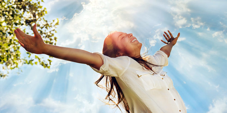 Pretty young woman raising her arms and enjoying perfect sunny day in nature. Her hair is long and bouncy, wearing a white shirt and enjoys smiling with eyes closed. Above her it is the sky with clouds through which spread sun rays. AdobeRGB color space. Small amount of grain added intentionally for better final impression.