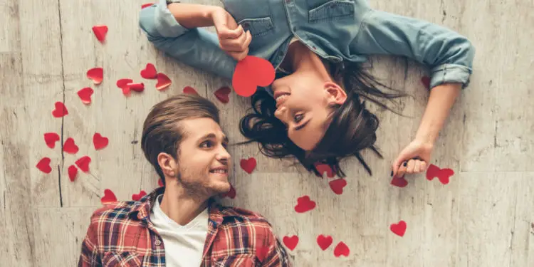 Top view of happy young couple looking at each other and smiling while lying on wooden floor. Girl is holding a red paper heart