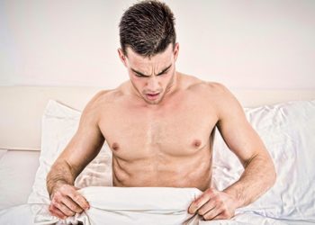 surprised half naked young man in bed  looking down at his underwear at his penis under white covers sheet in badroom. Concept photo of male sexuality and man sex problems, domestic atmosphere.