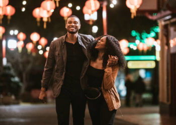 A smiling African American man and woman walk arm in arm on the streets of Chinatown in L.A. California on a warm evening, exploring the cities night life.  Bright traditional lanterns illuminate the scene.