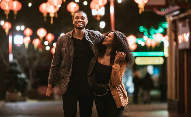 A smiling African American man and woman walk arm in arm on the streets of Chinatown in L.A. California on a warm evening, exploring the cities night life.  Bright traditional lanterns illuminate the scene.