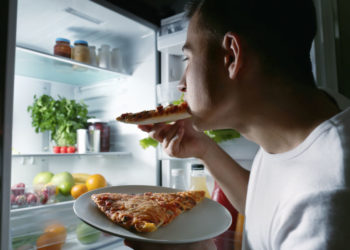 Young man eating pizza near refrigerator at night