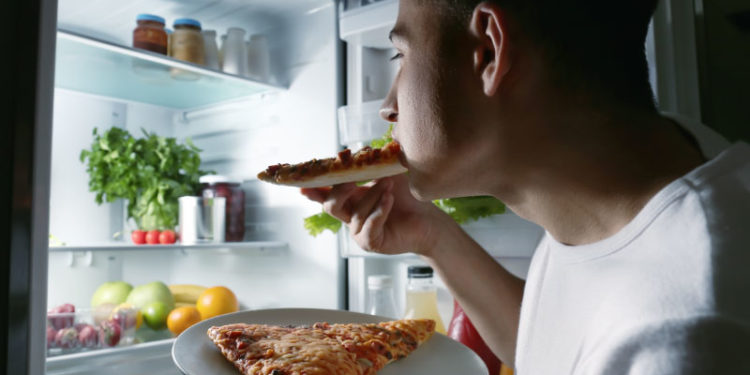 Young man eating pizza near refrigerator at night
